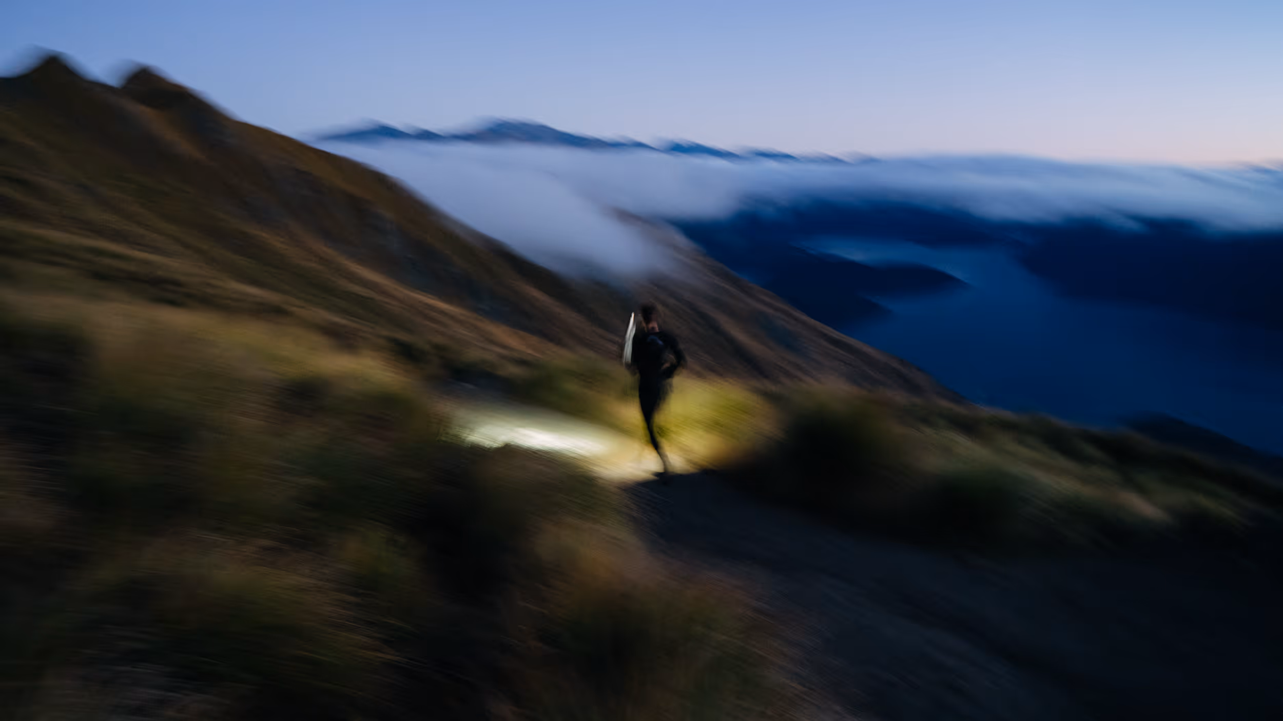 Blurred image of a person running on a mountain trail at dusk with clouds over distant hills.