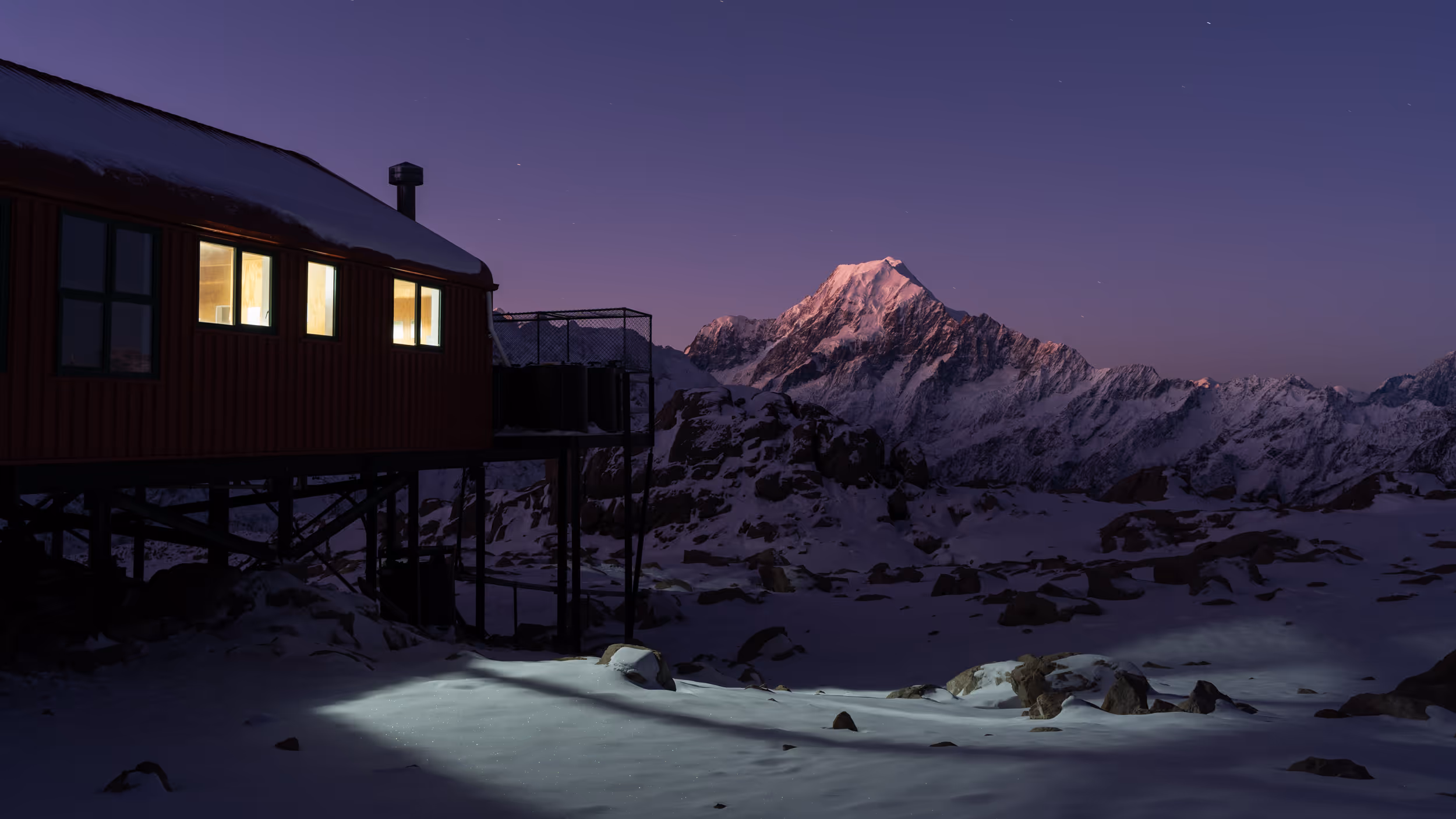 Mountain cabin with lit windows overlooking snowy terrain at dusk with snow-covered peak in the distance.