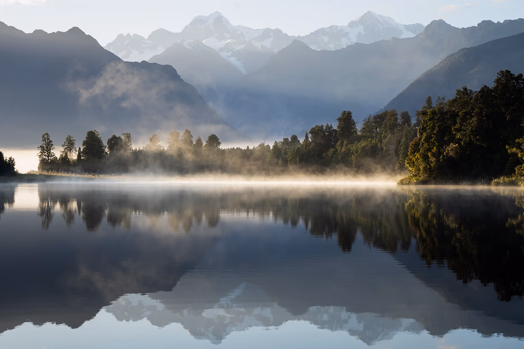 Calm lake reflecting mist-covered trees and snow-capped mountains in the background.
