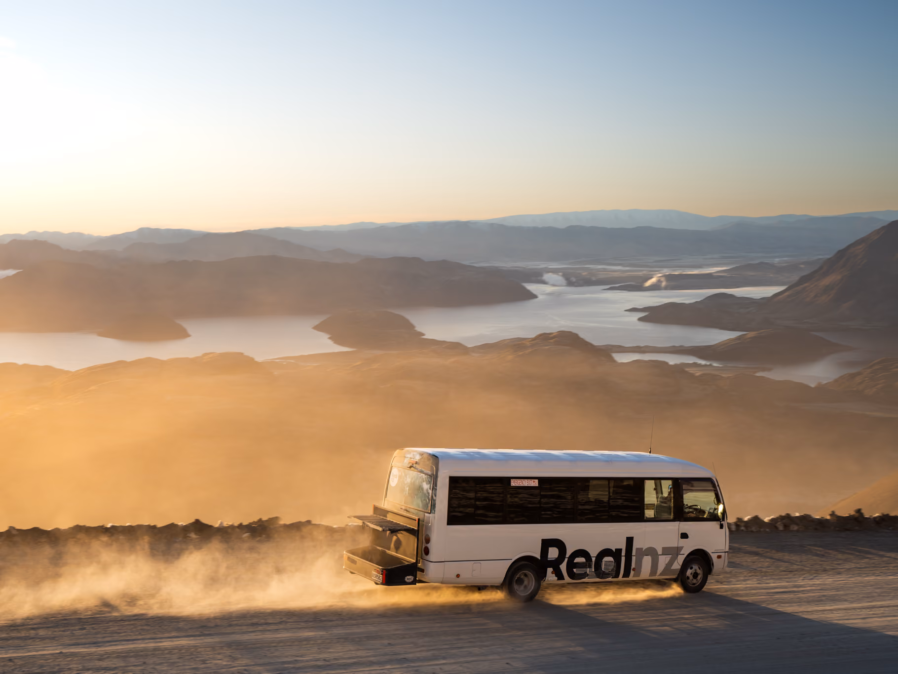 White bus with 'Realnz' branding driving on a dusty mountain road overlooking a lake and distant hills at sunset.