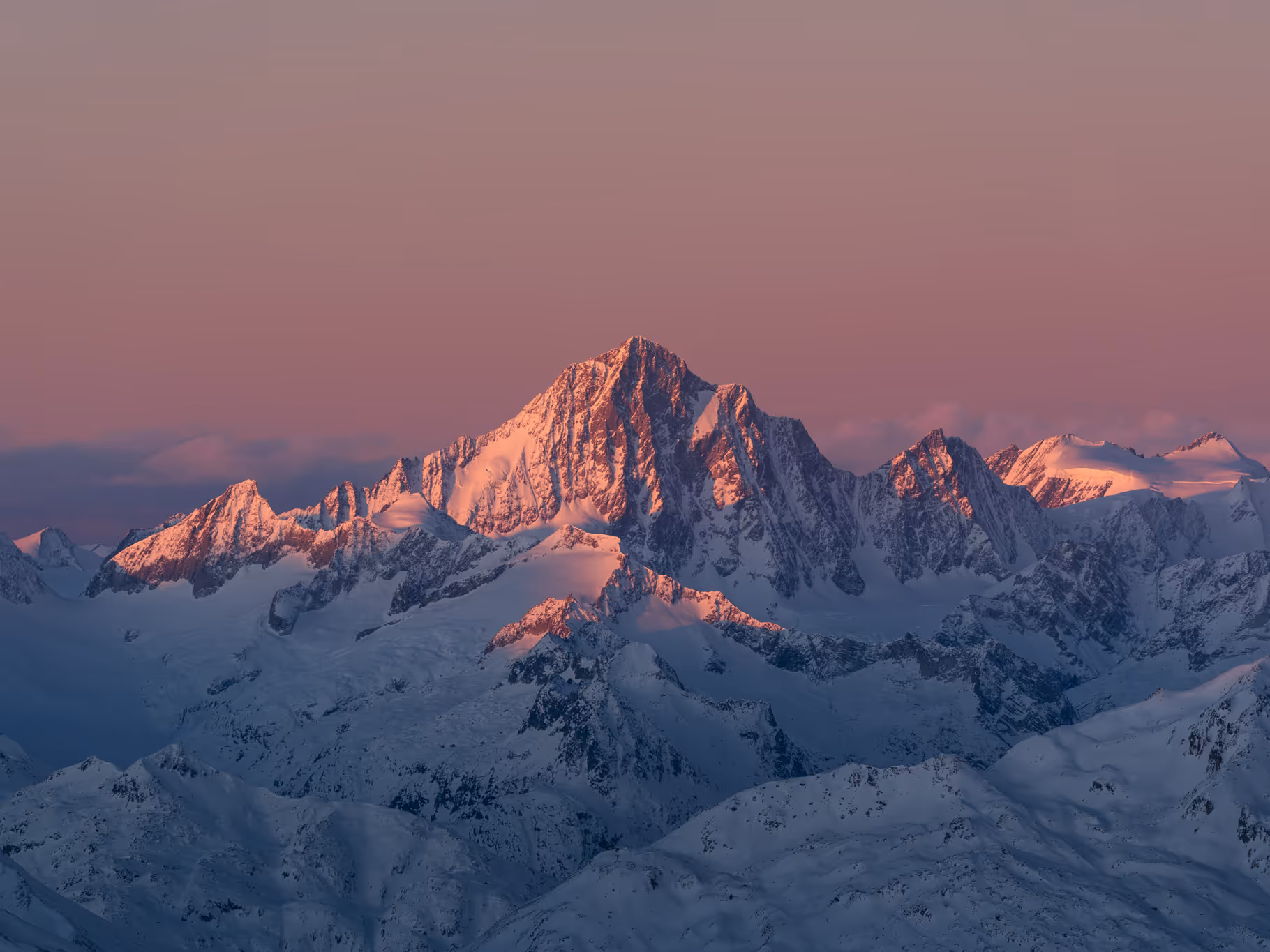 Snow-covered mountain peaks illuminated by soft pink and purple light during sunrise or sunset.