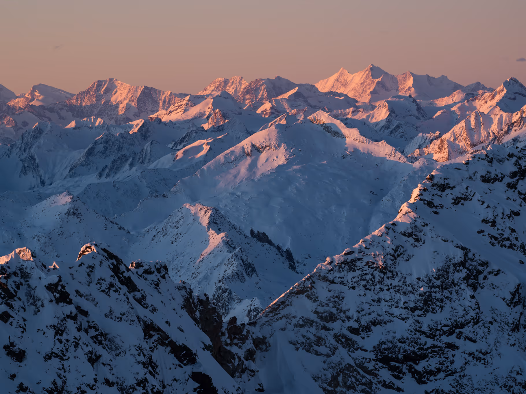 Mountain range covered in snow illuminated by the soft pink light of sunrise or sunset.