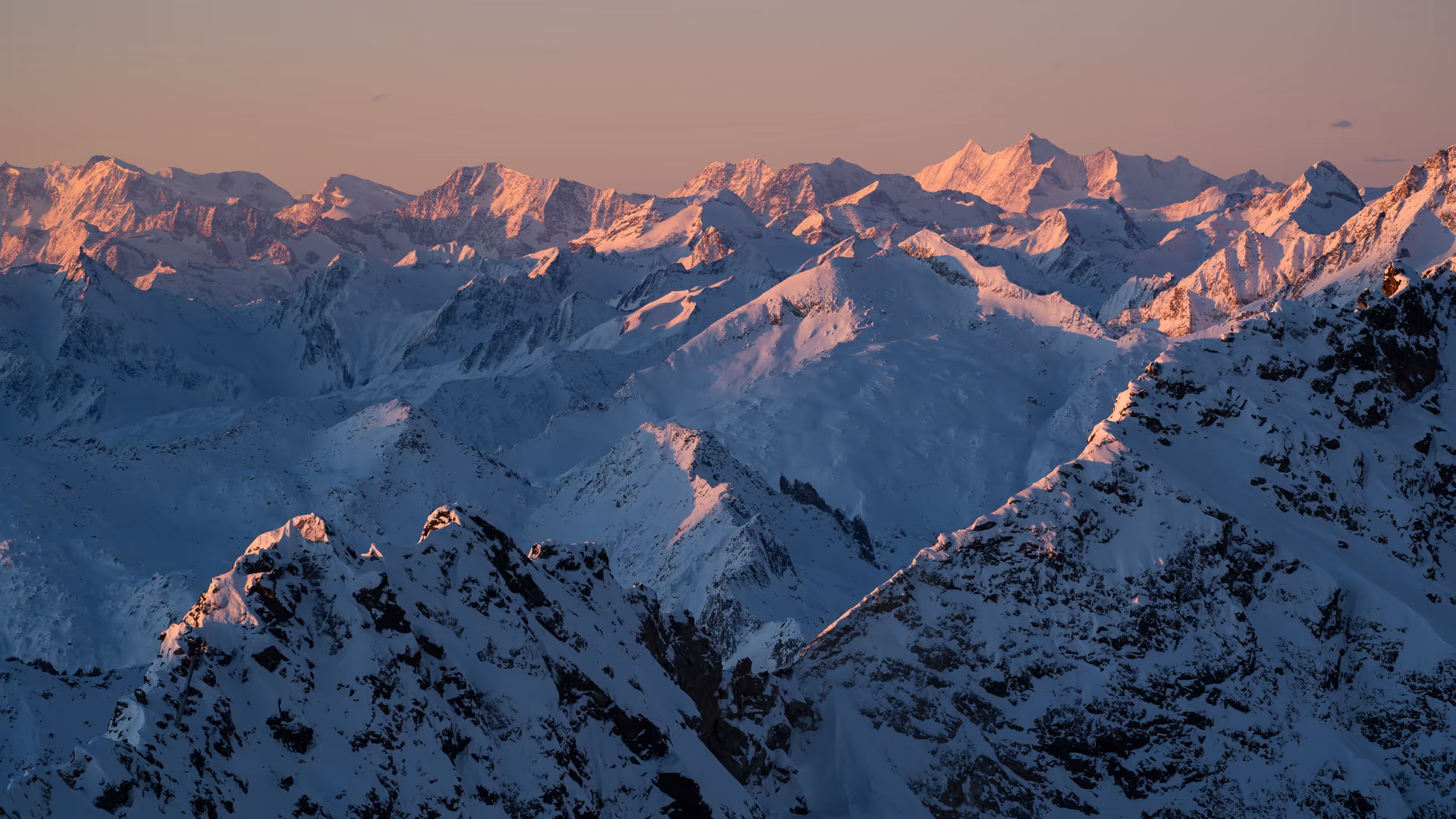Snow-covered mountain peaks illuminated by soft pink light at sunrise or sunset.