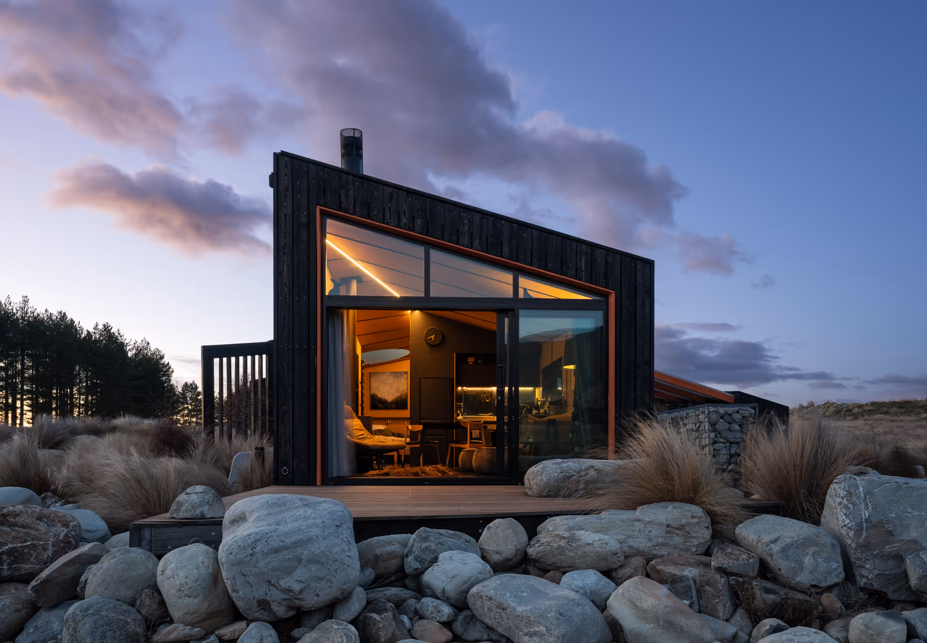 Modern small cabin with large glass doors lit warmly inside, surrounded by rocks and tall grasses at dusk.