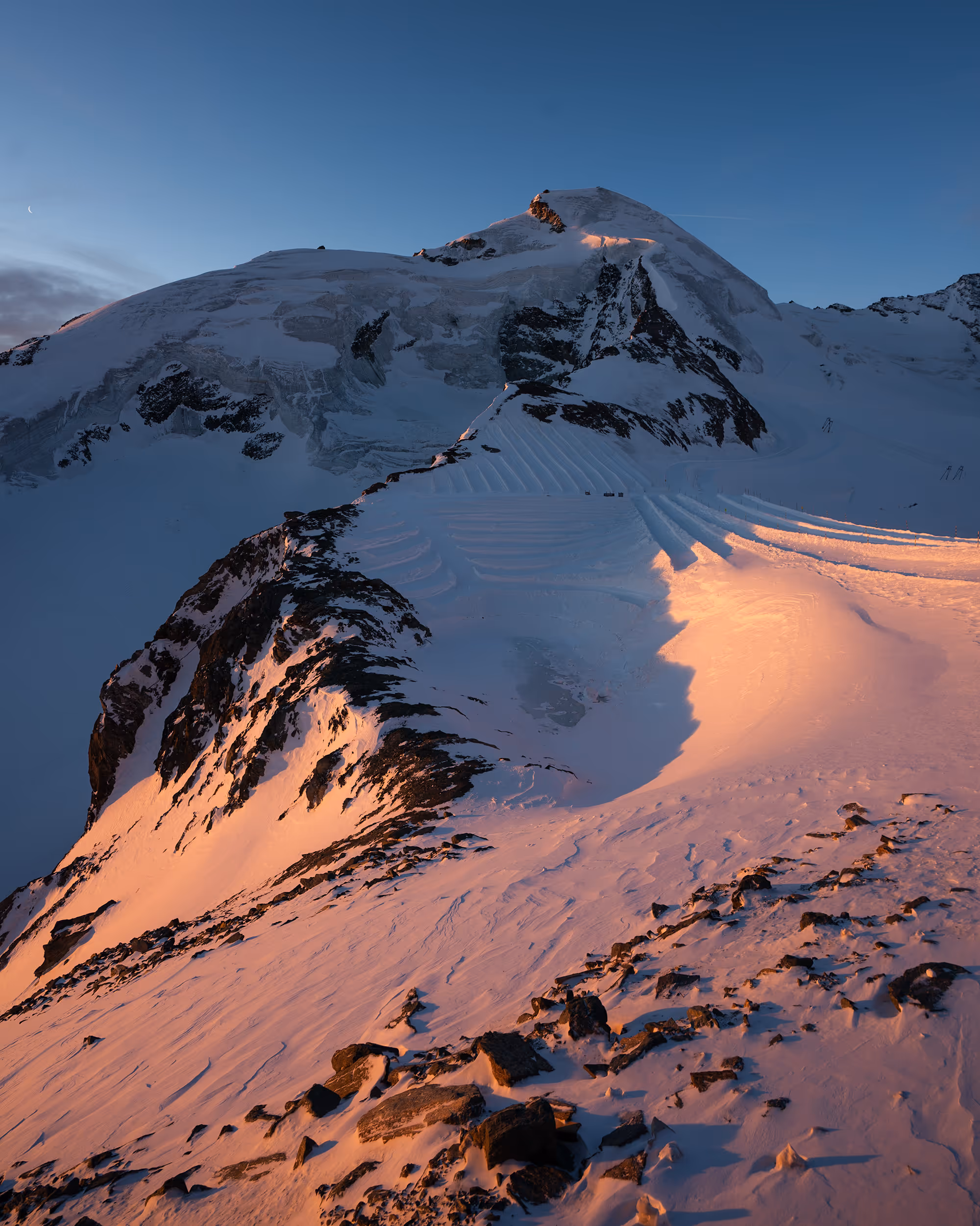 Snow-covered mountain peak in warm sunrise light with rocky foreground under clear blue sky.
