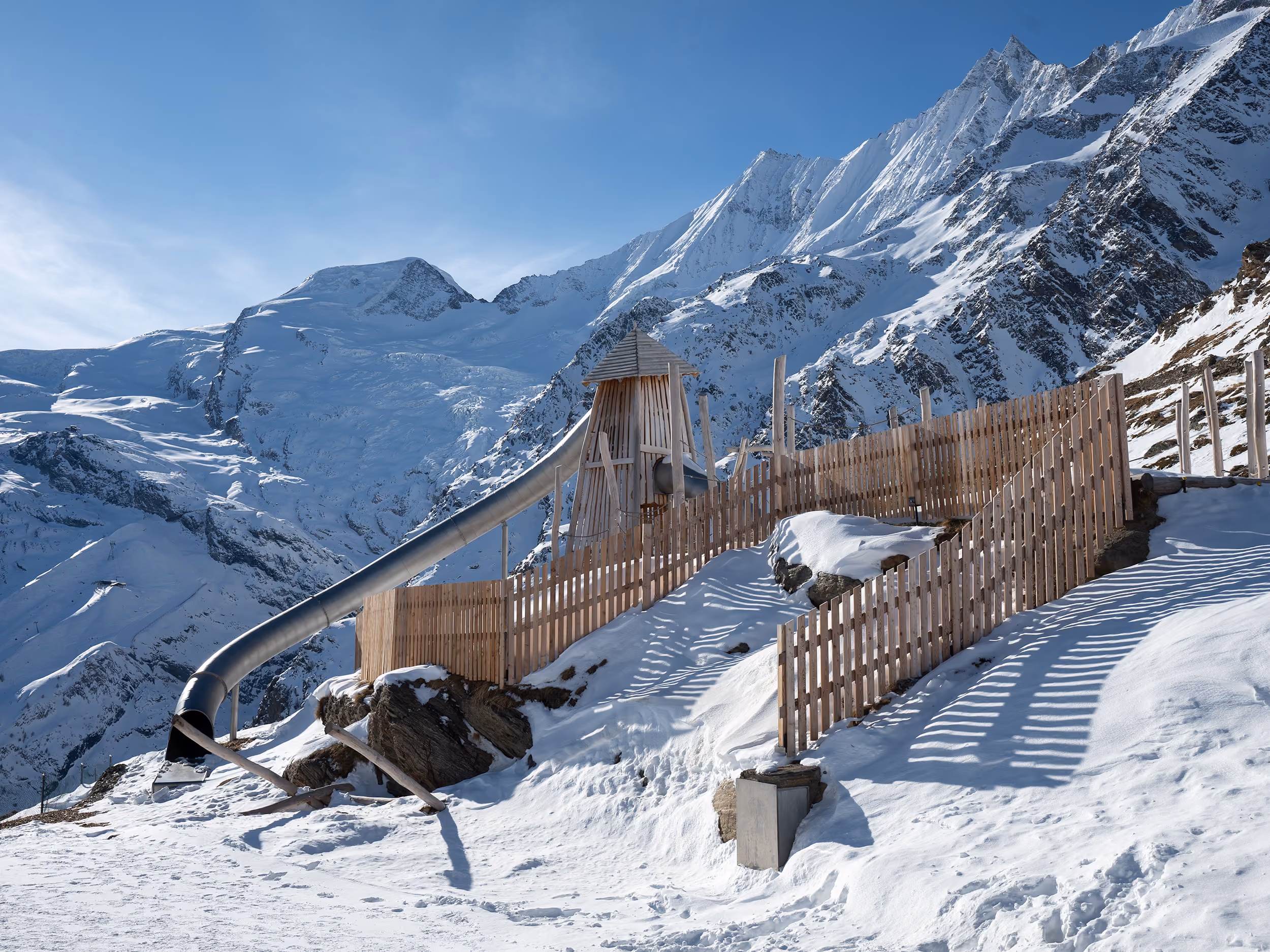 Snow-covered mountain playground with a tall wooden slide and fenced area under clear blue sky.