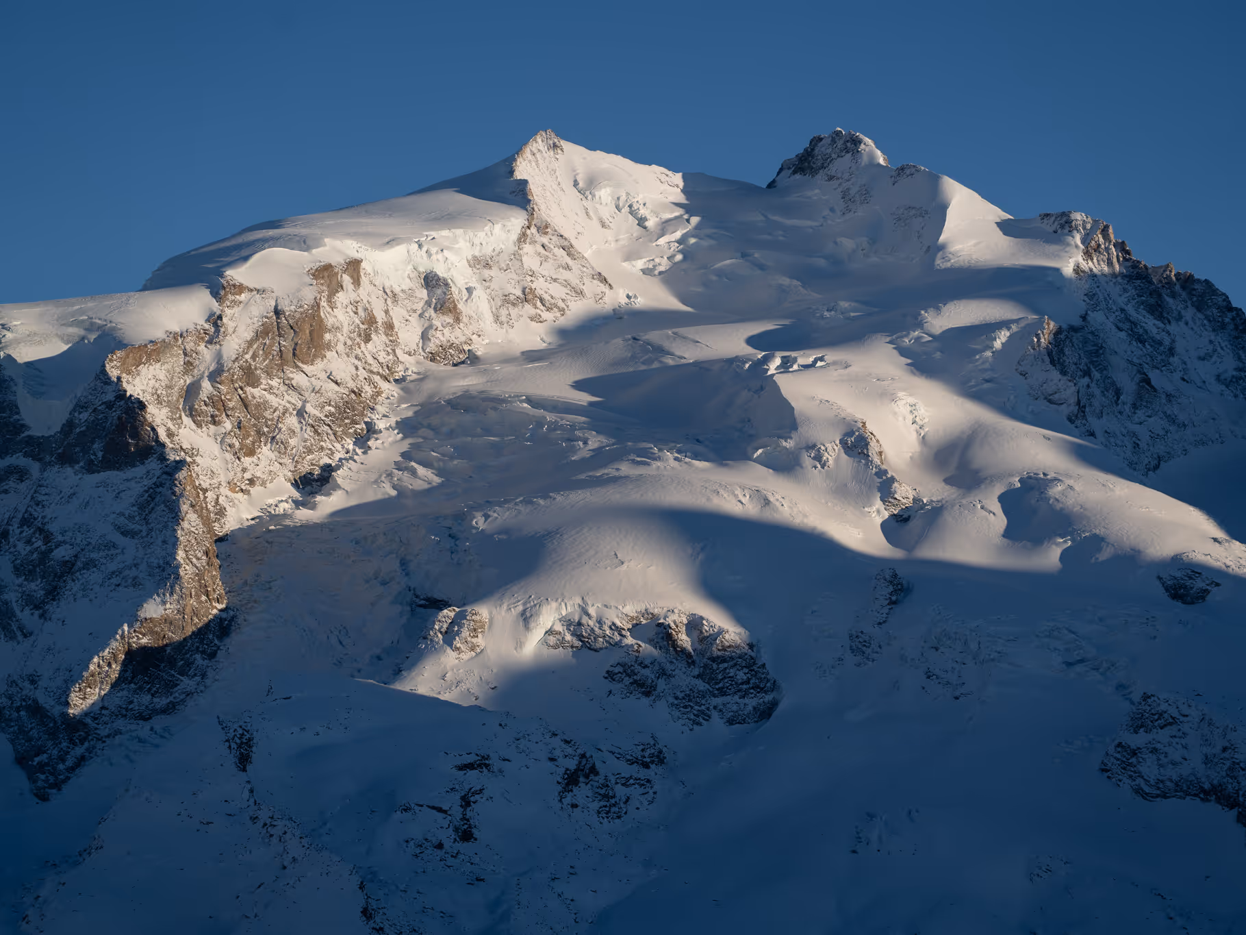 Snow-covered mountain peak under clear blue sky with shadows on the slopes.