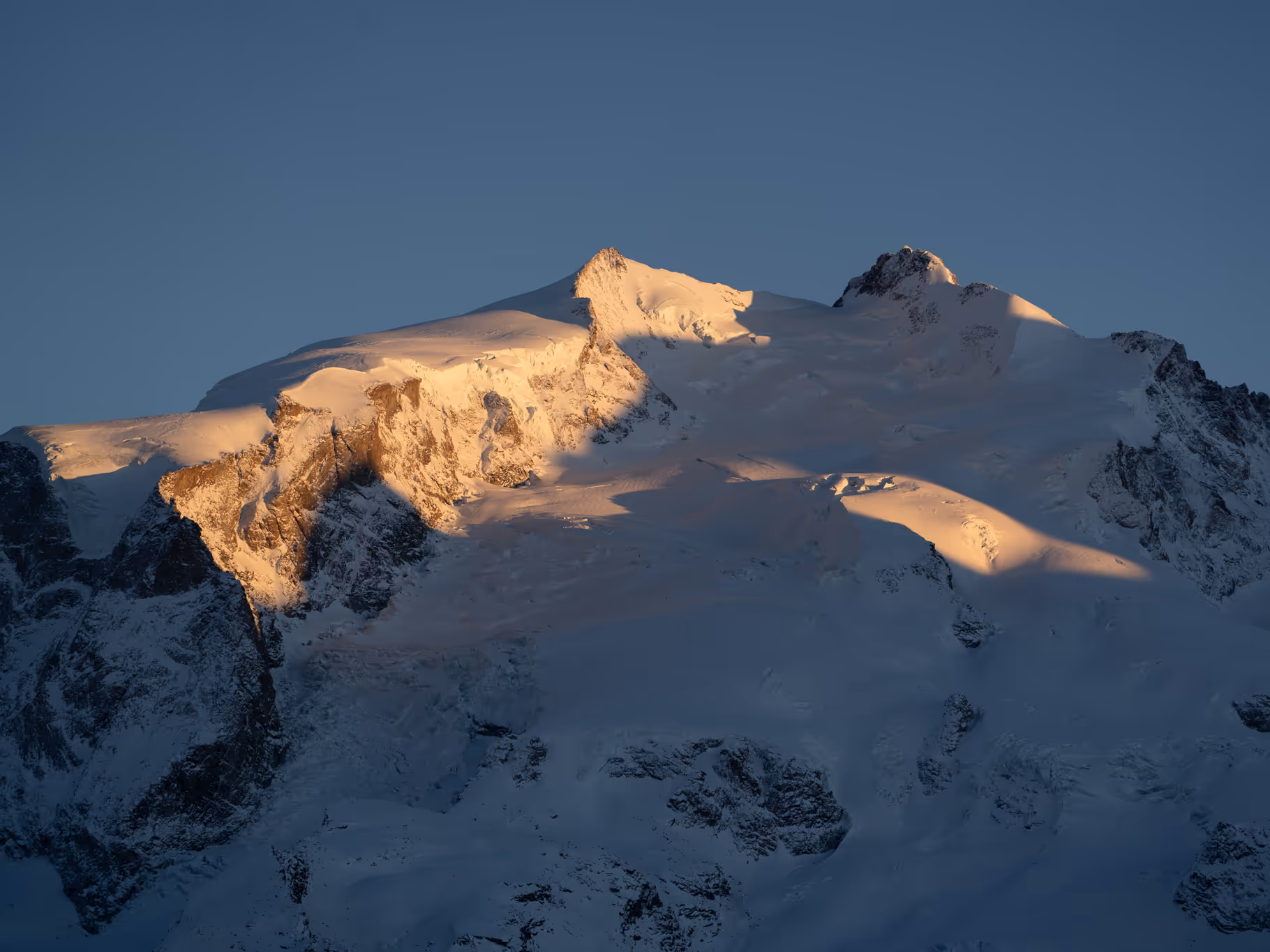 Snow-covered mountain peaks illuminated by golden sunlight against a clear blue sky.