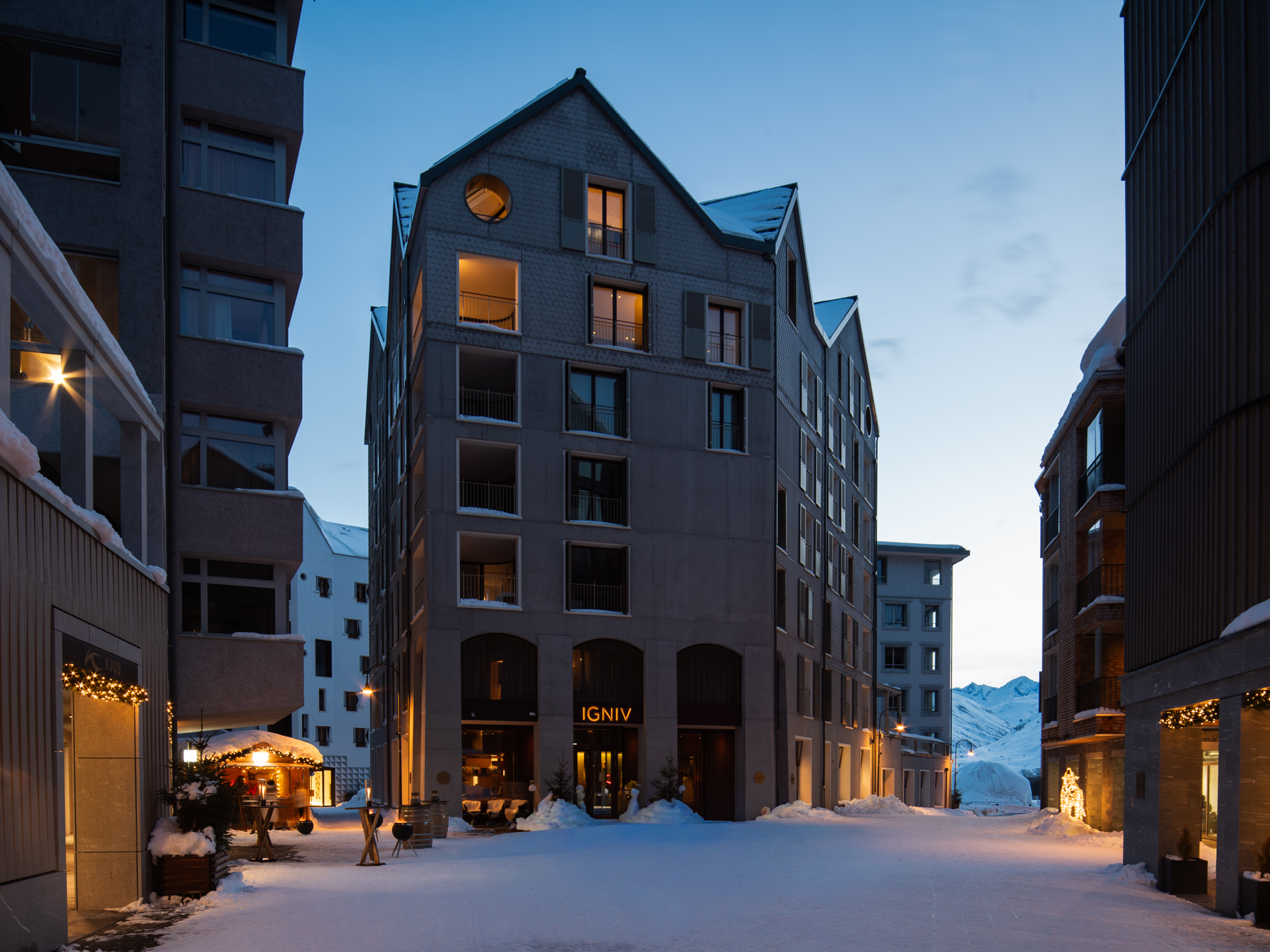 Snow-covered street lined with modern buildings at dusk with lights on and mountains visible in the background.