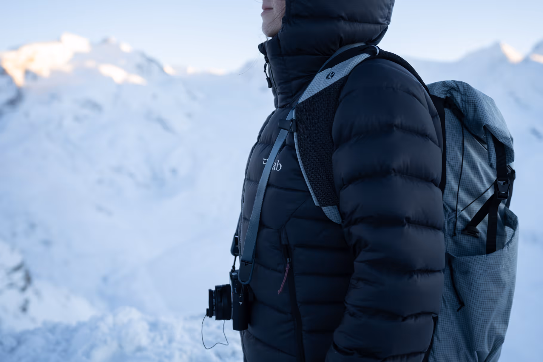 Person wearing a black puffer jacket with a hood and carrying a grey backpack in a snowy mountain environment.