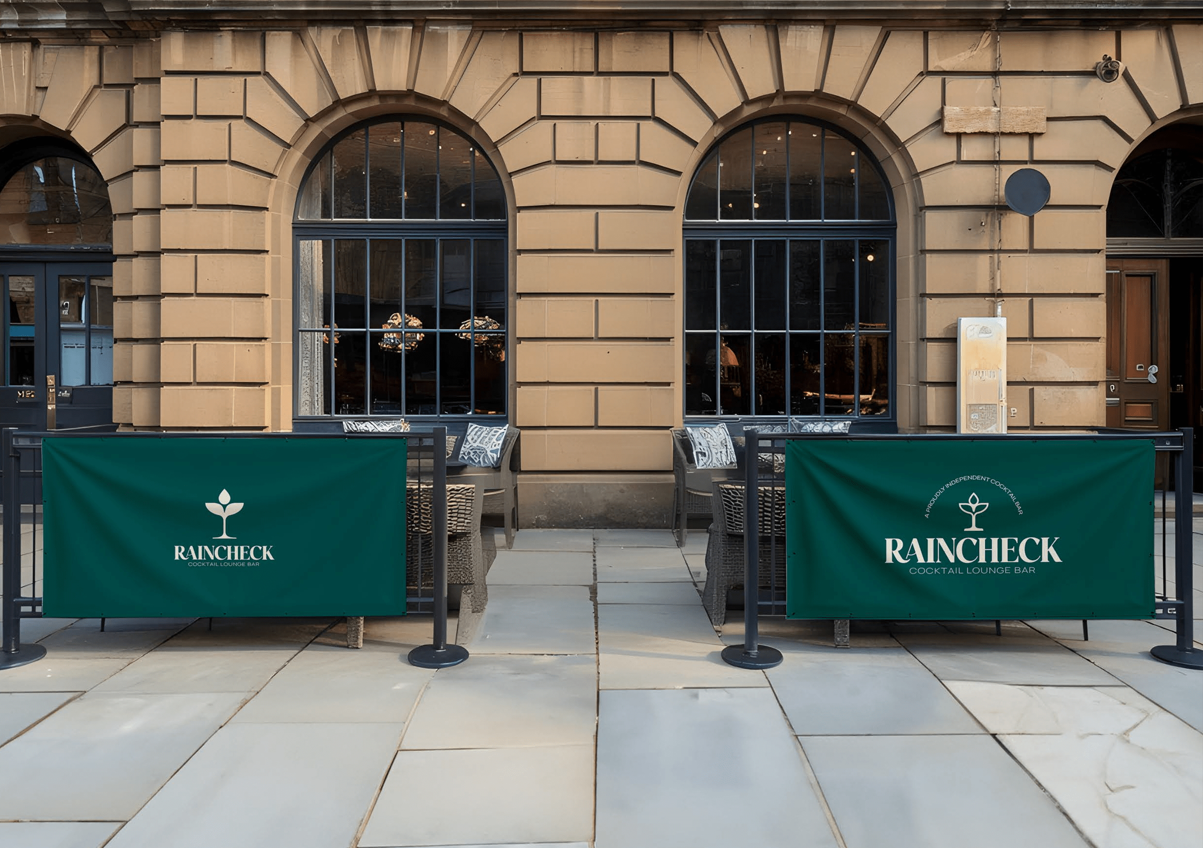Outdoor seating area with wicker chairs and green barriers displaying Raincheck Cocktail Lounge Bar logo in front of arched windows on a beige brick building.