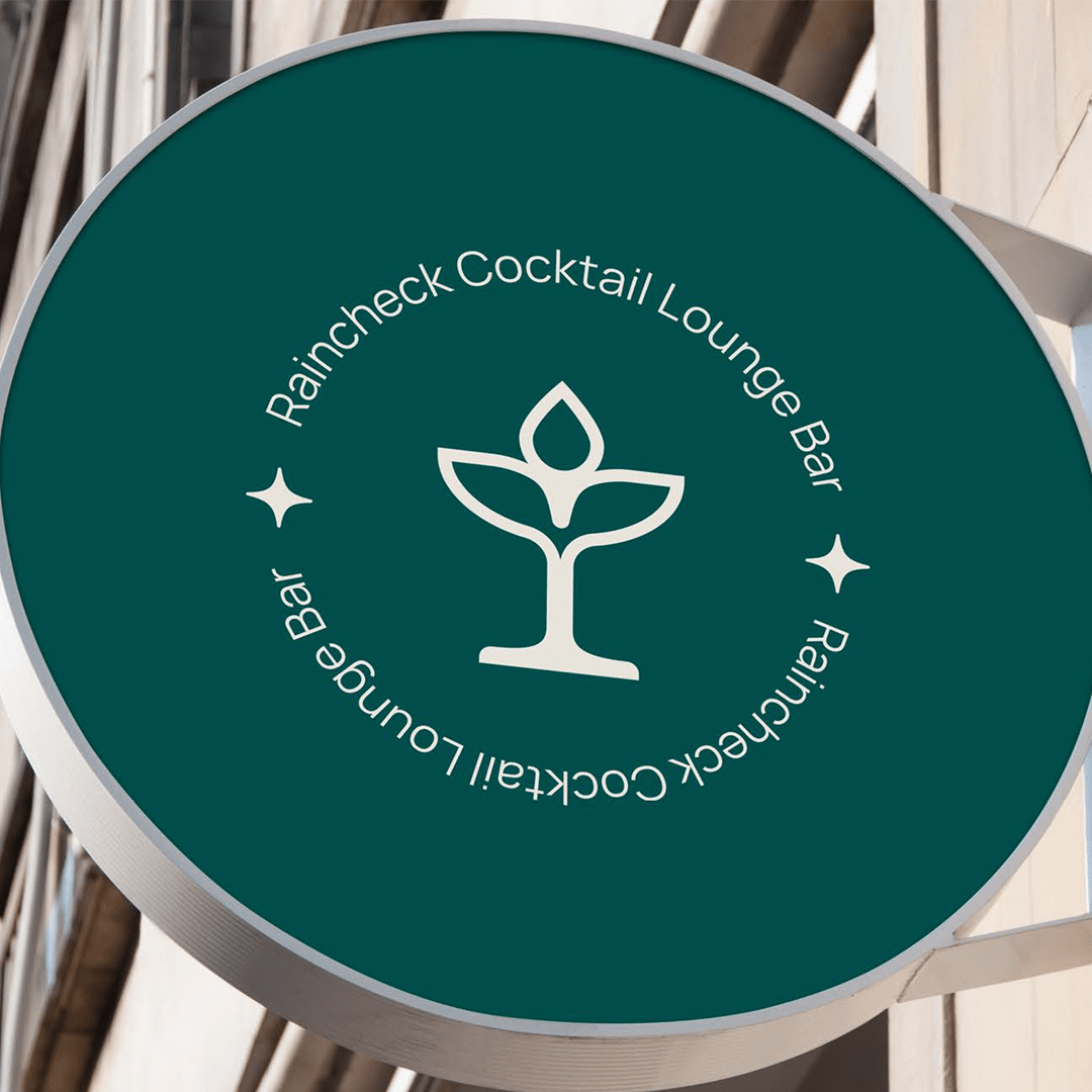 Round green sign for Raincheck Cocktail Lounge Bar with a stylized cocktail glass and leaf logo in white.