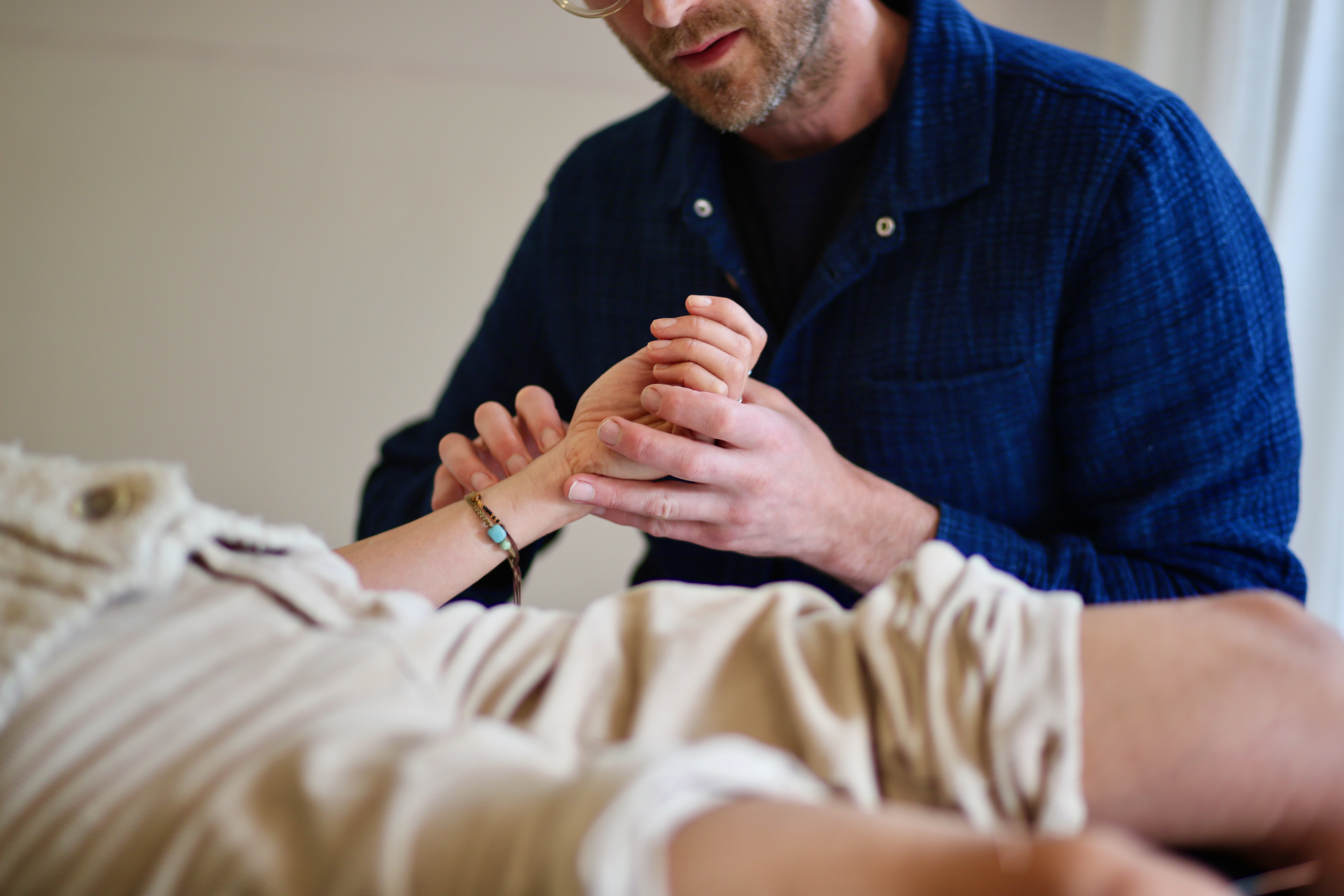 Man in blue shirt examining wrist of a person lying down during a wellness session.