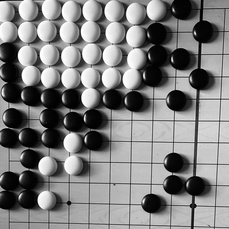 Black and white stones placed on a Go board, showing an ongoing game with a dense cluster of white stones surrounded by black stones on the upper left and a few scattered stones on the right side.