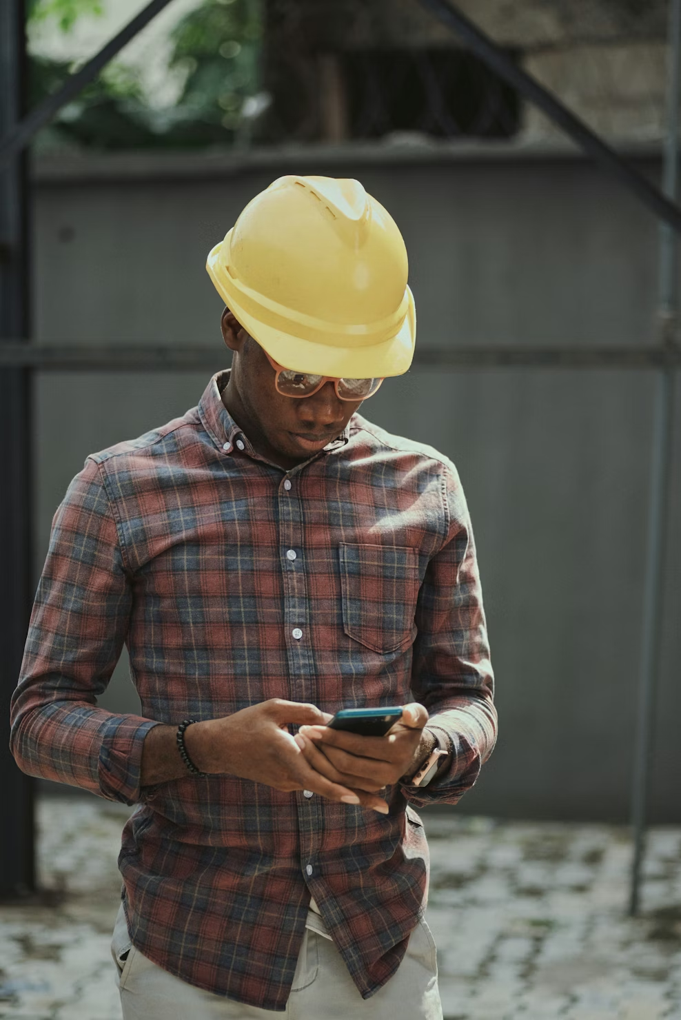 Man on construction site wearing a hard hat, looking down at smartphone. 