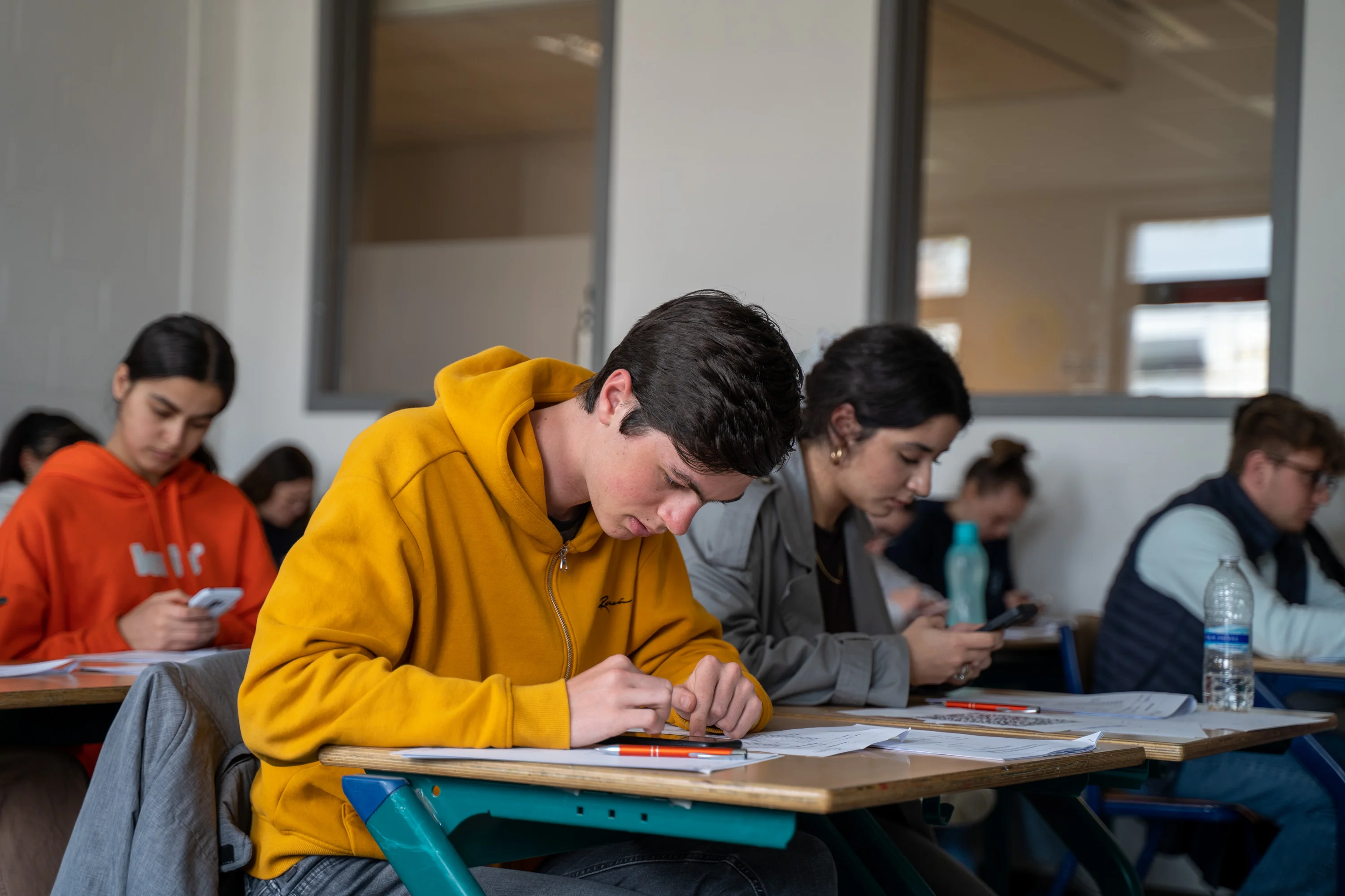 Des étudiants concentrés en classe, certains écrivent sur papier et d'autres utilisent leur téléphone.