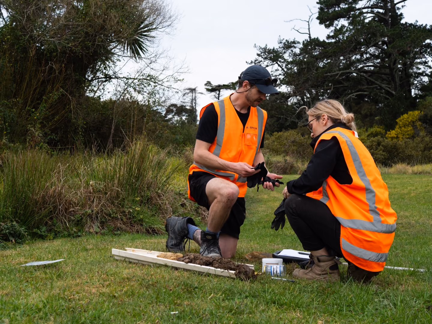 Two on-site qualified environmental scientists wearing high-vis vests and hard hatsassessing ongoing earthworks near a freshwater waterway for contamination in an active construction site