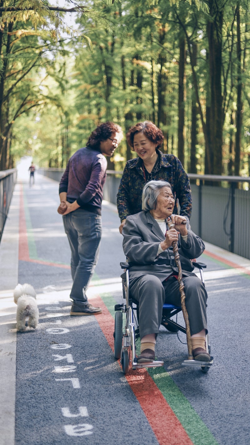 Family members walking with elderly loved one in wheelchair through a park, showing the support families provide