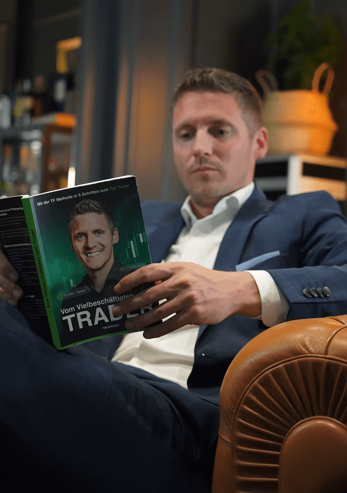 Mann in Anzug liest Buch über Trading auf einem Ledersessel, Bücherregal im Hintergrund.