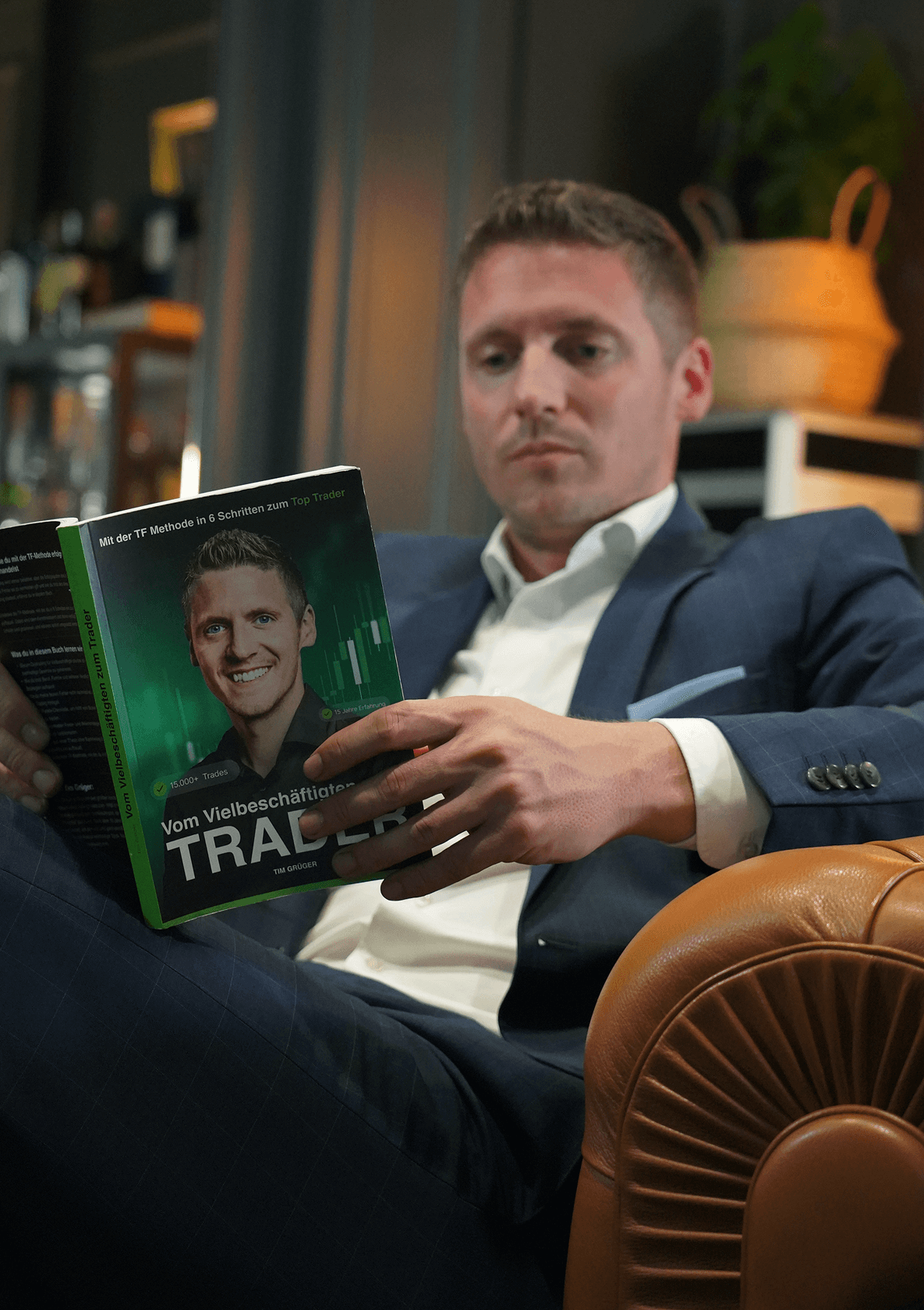 Mann in Anzug liest Buch über Trading auf einem Ledersessel, Bücherregal im Hintergrund.