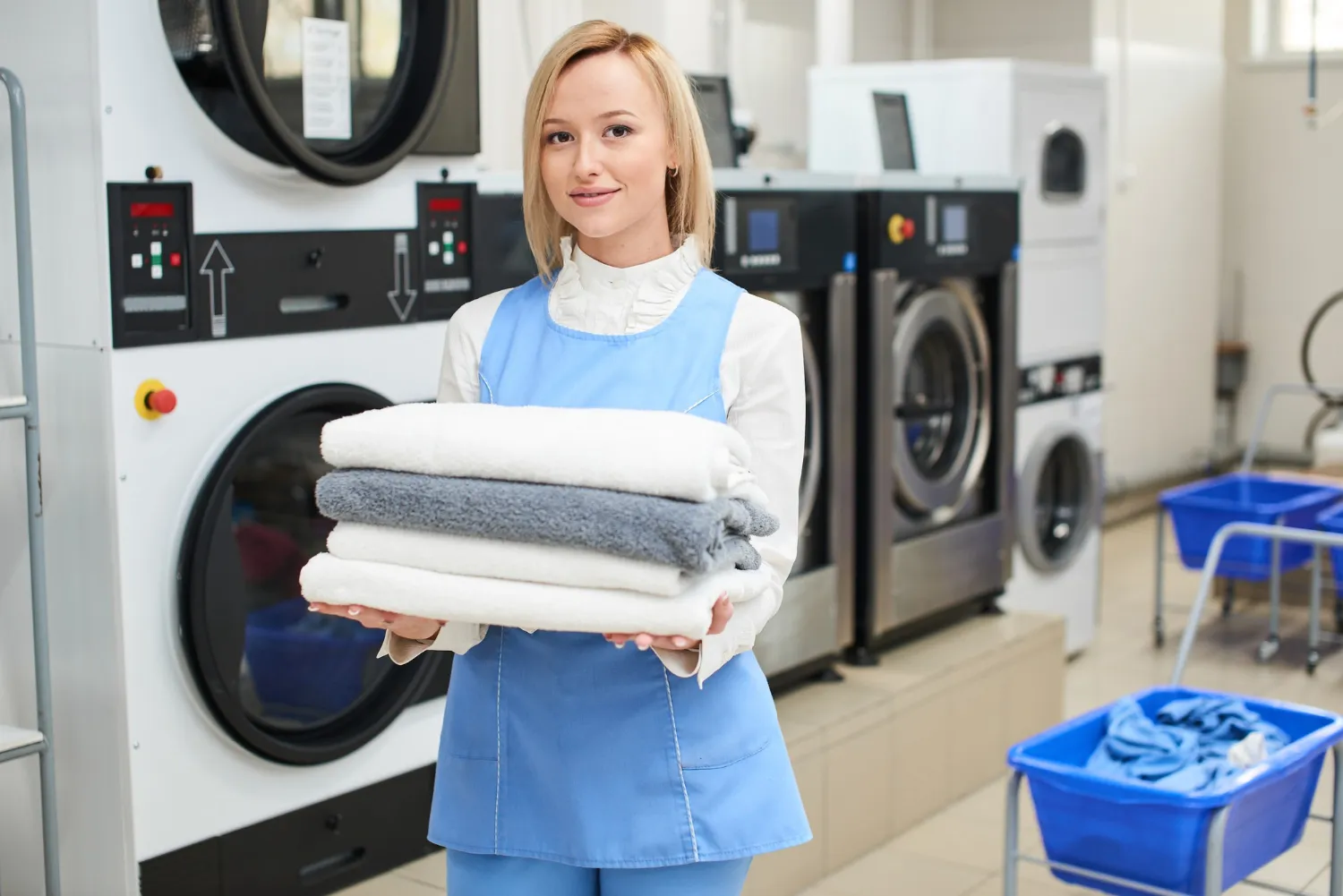 Laundry worker in blue uniform holding a stack of folded towels in a commercial laundry room.