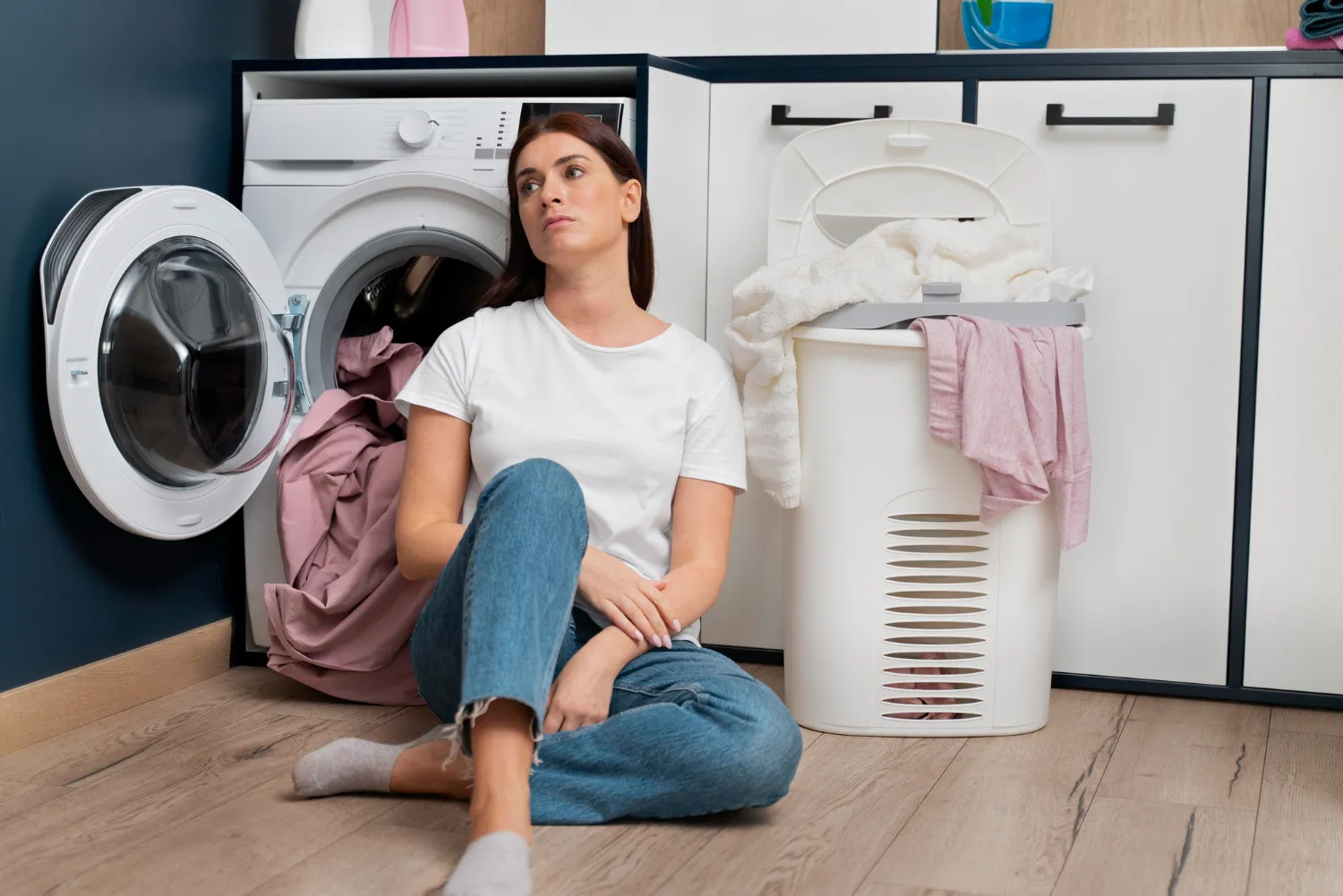 Woman in white t-shirt and jeans sitting on the floor next to a washing machine with its door open and a laundry basket filled with clothes.