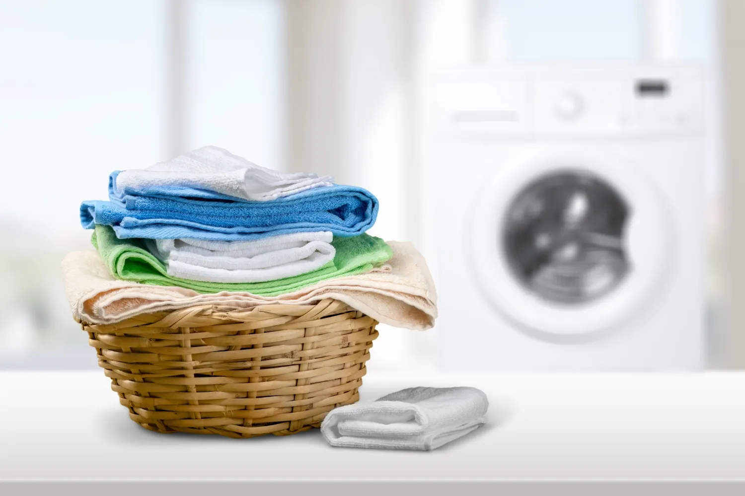 Wicker basket filled with neatly folded colorful towels next to a folded white towel, with a washing machine in the background.
