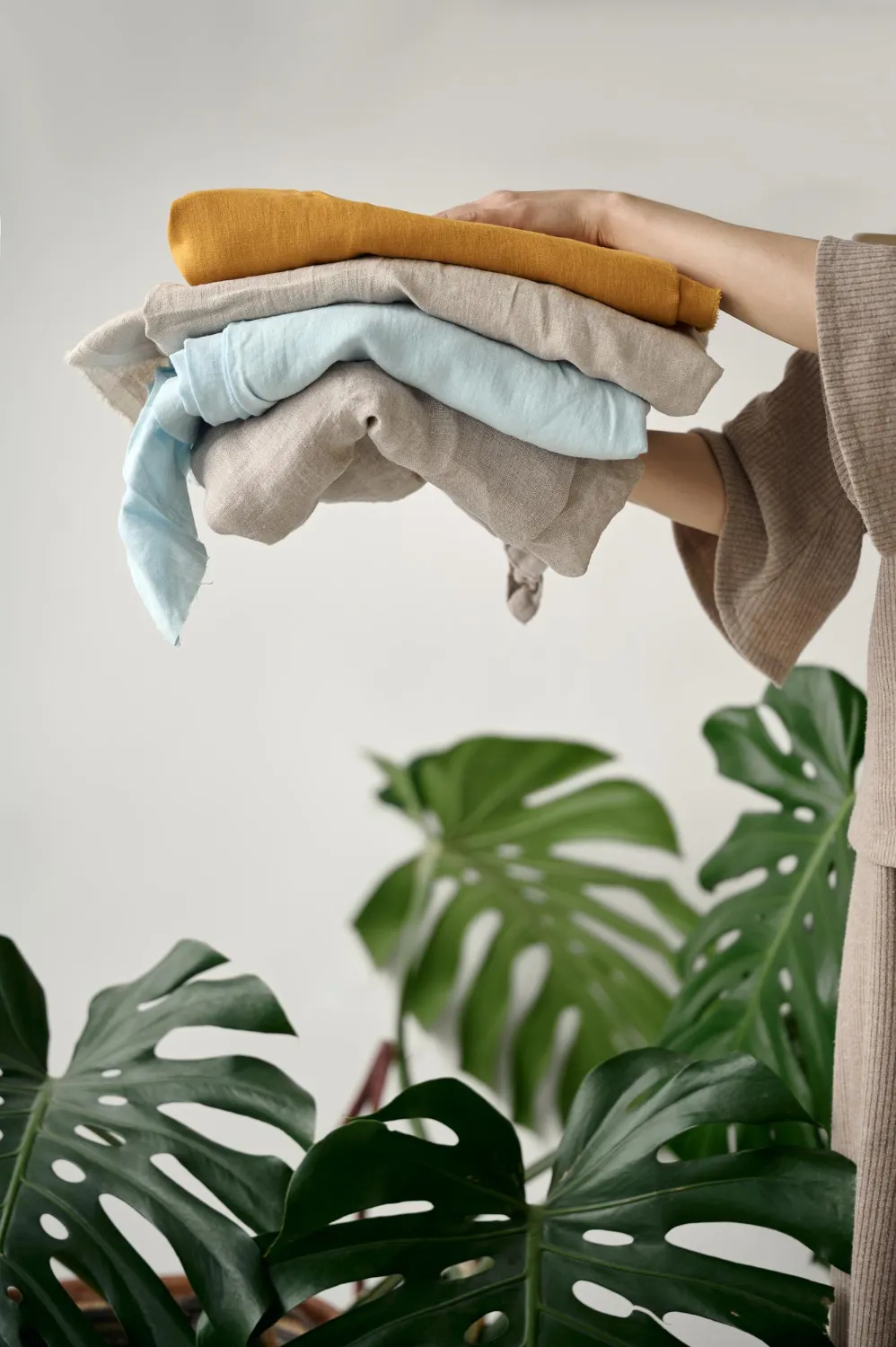 Person holding a stack of folded linen clothes in beige, blue, and mustard colors with green monstera leaves in the background.