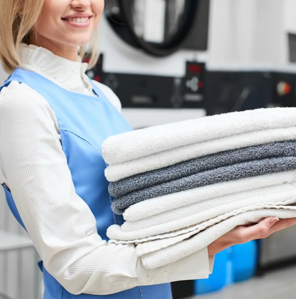 Smiling woman in a blue apron holding a stack of clean white and gray towels inside a laundry facility.