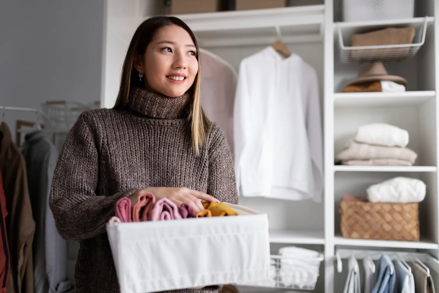 Smiling woman holding a white basket with neatly folded colorful clothes in a closet.