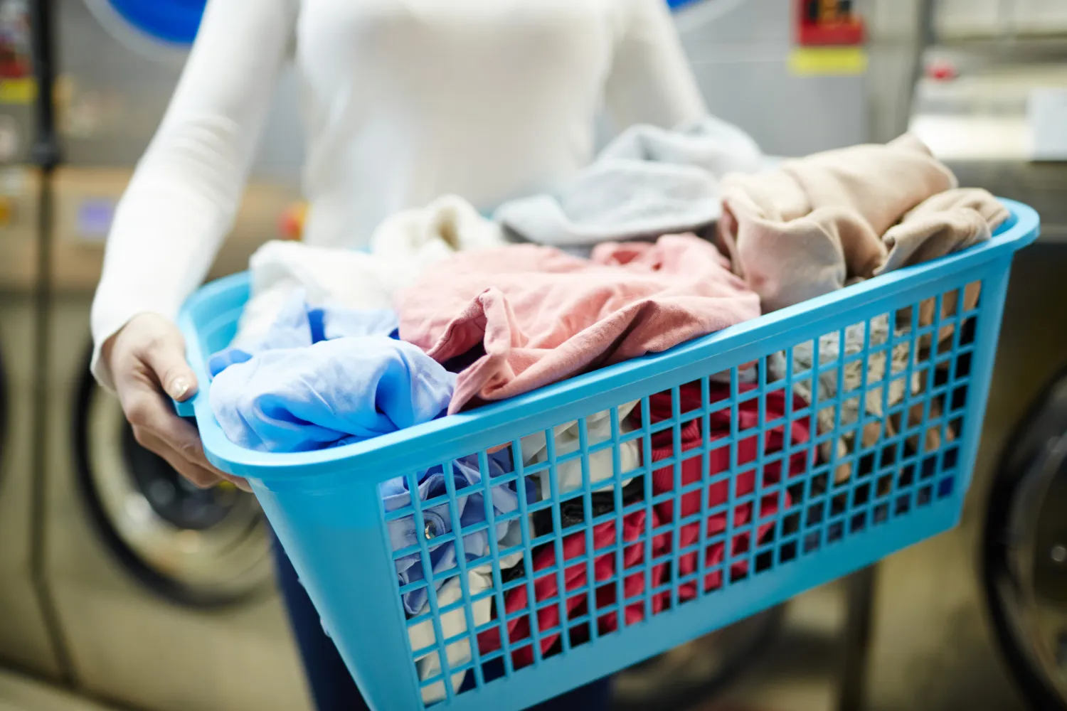 Person holding a blue laundry basket filled with assorted clean clothes in a laundry room.