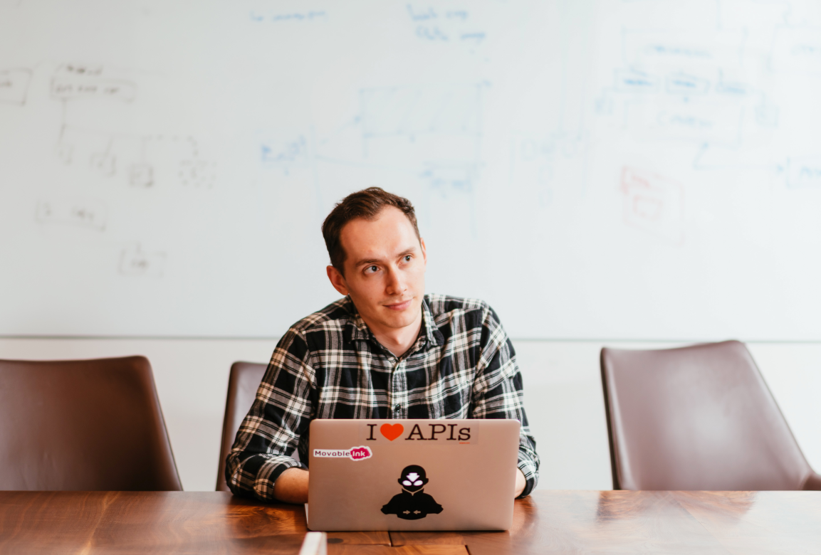 Young man in a plaid shirt sitting at a conference table looking thoughtfully past his laptop with API-themed stickers.