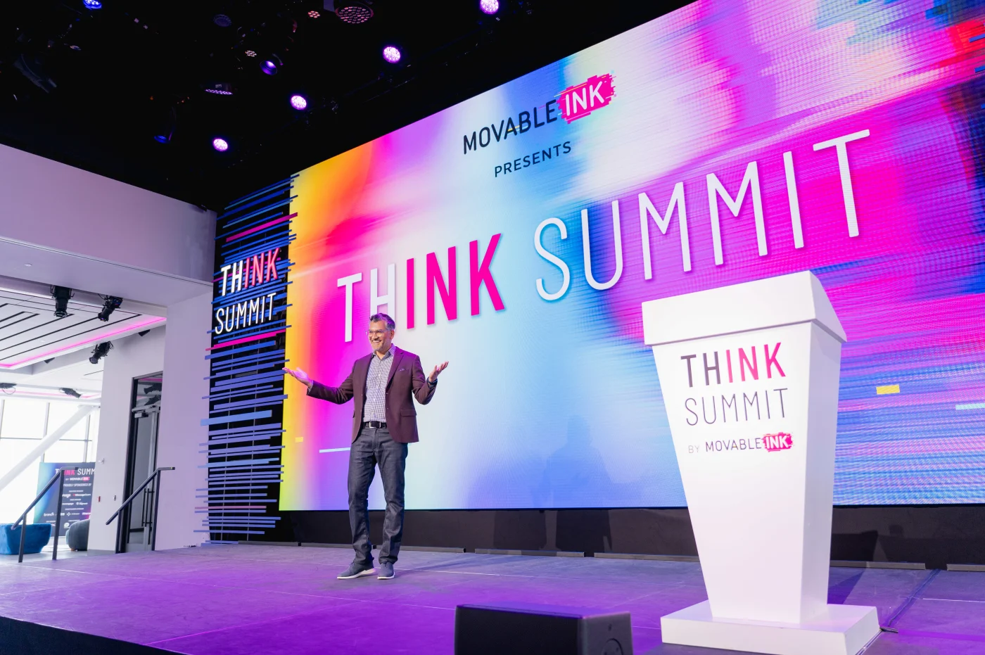 Man standing on stage speaking at THINK SUMMIT event with colorful background screen and white podium.