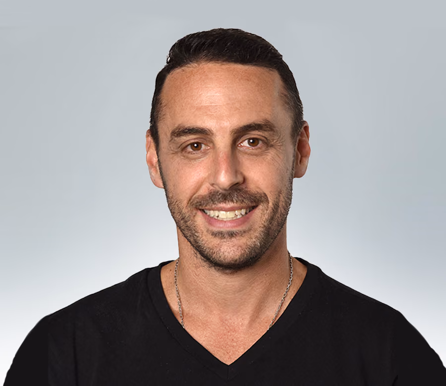 Smiling man with short dark hair and a beard wearing a black V-neck shirt and silver necklace against a light gray background.
