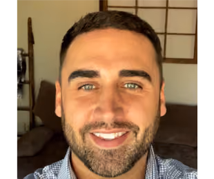 Smiling man with short dark hair and beard wearing a checkered shirt in a room with brown furniture and a window with wooden frame.