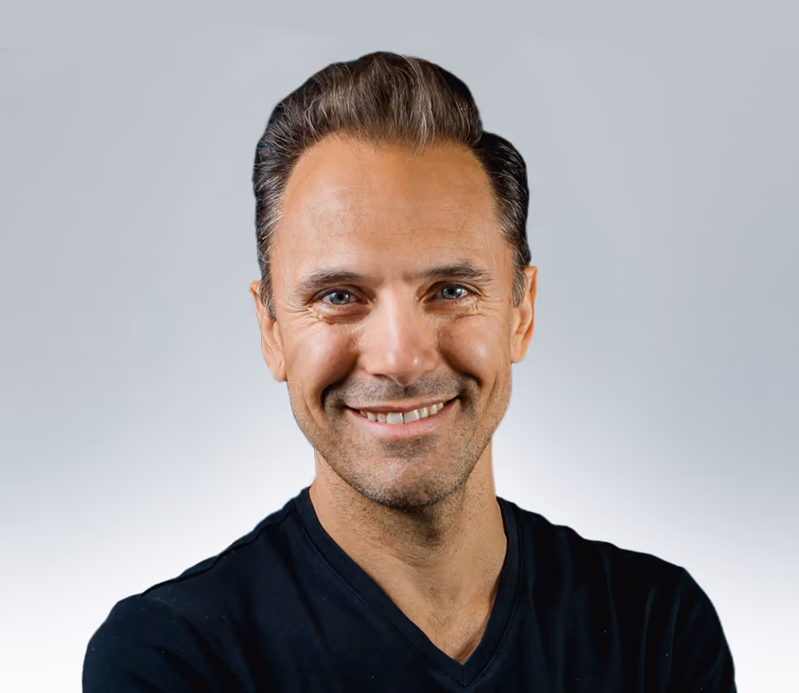 Smiling middle-aged man with short dark hair wearing a black V-neck shirt against a plain light gray background.