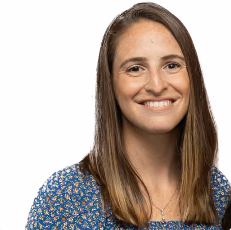 Smiling woman with long straight brown hair wearing a blue floral top and a small pendant necklace against a white background.