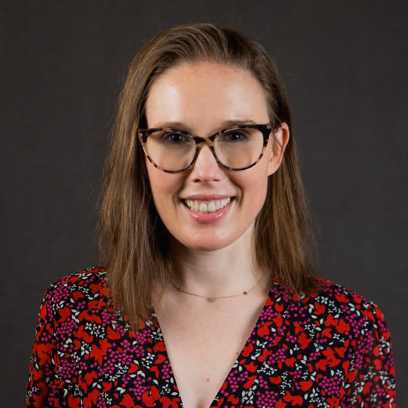 Smiling woman with straight brown hair, wearing tortoiseshell glasses and a red floral patterned blouse against a dark background.