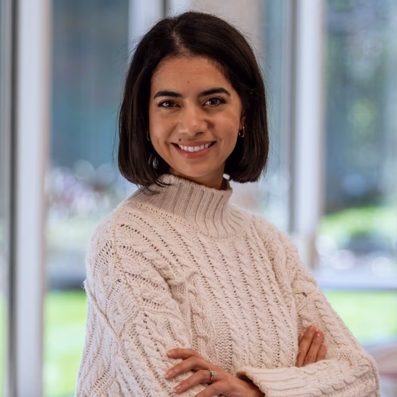 Smiling woman with dark hair wearing a cream cable-knit sweater standing indoors with arms crossed.