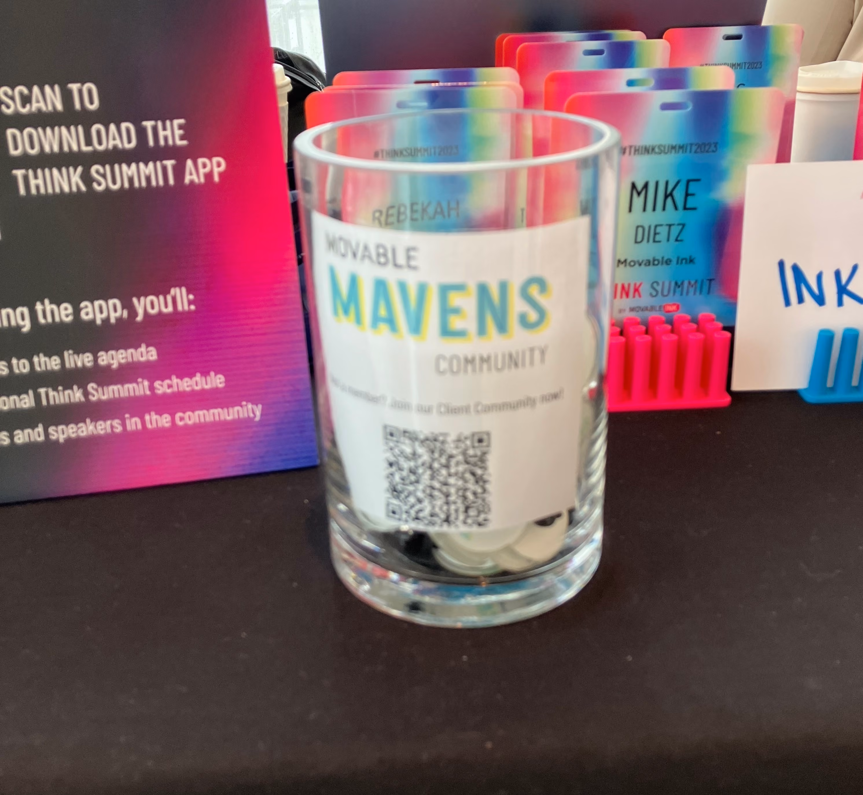 Glass container labeled Movable Mavens Community holding round tokens on a table with colorful name badges and a sign promoting the Think Summit app in the background.