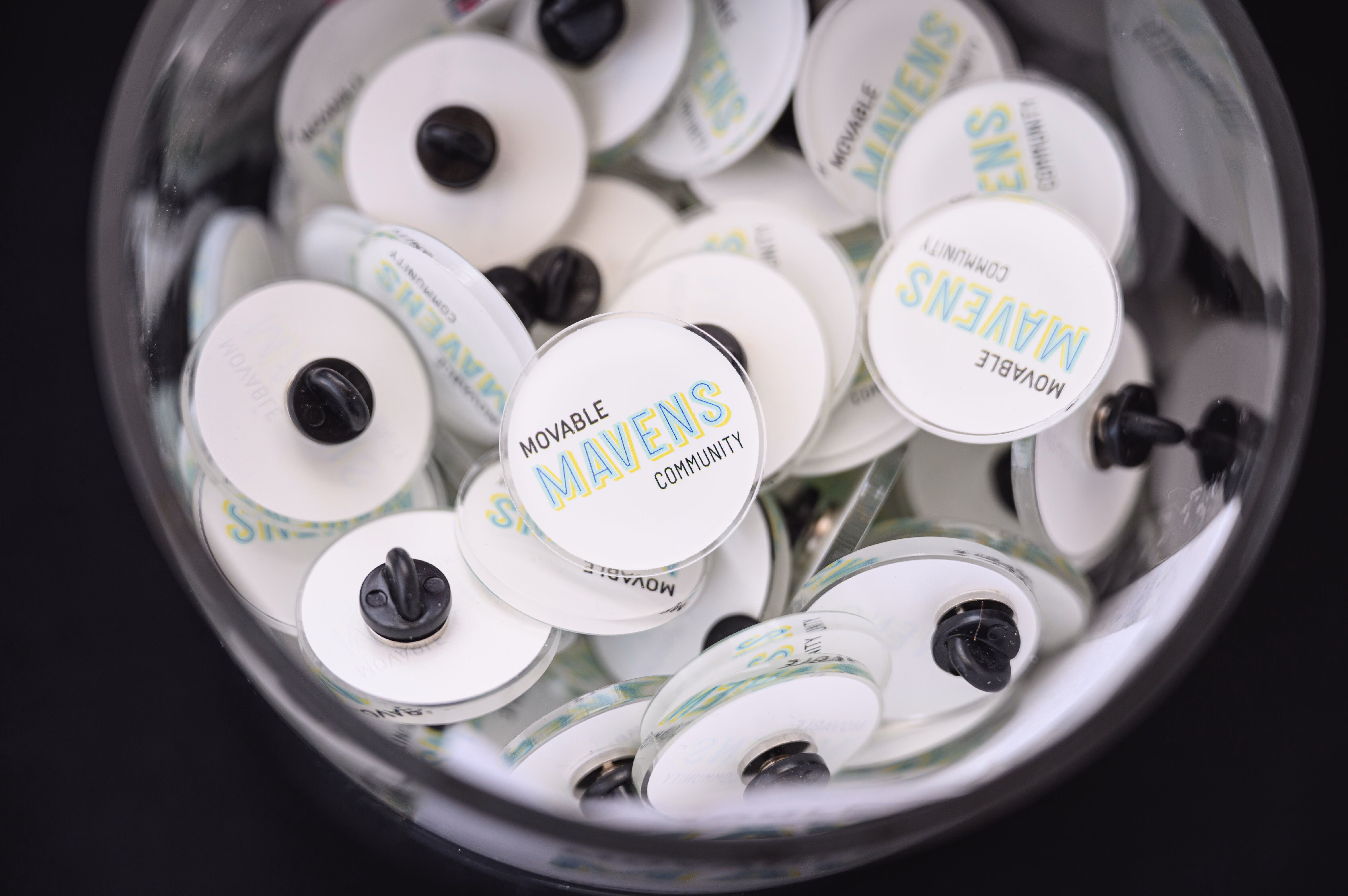 Close-up of a glass bowl filled with round white pins labeled 'Movable Mavens Community' with black pin backs.