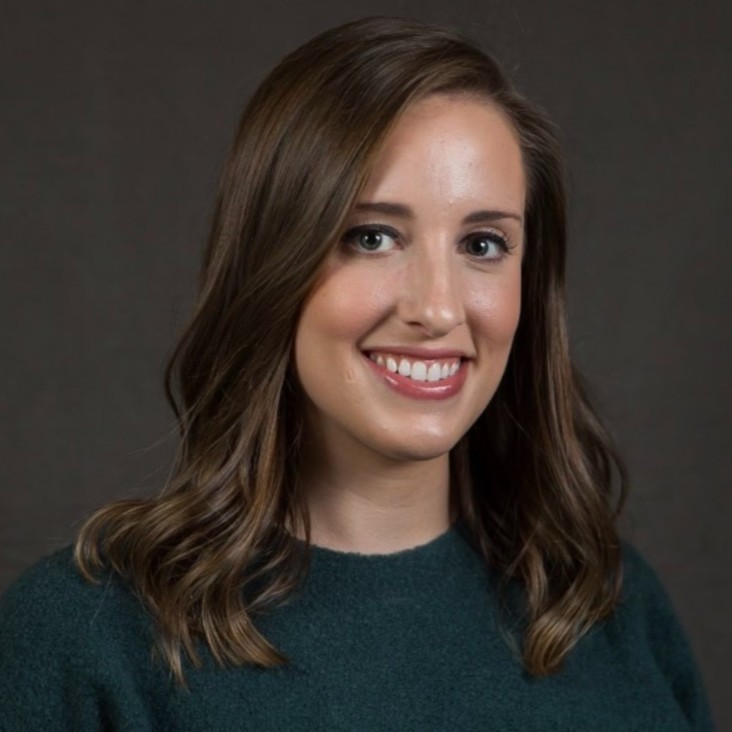 Smiling woman with shoulder-length brown hair wearing a dark green sweater against a dark background.