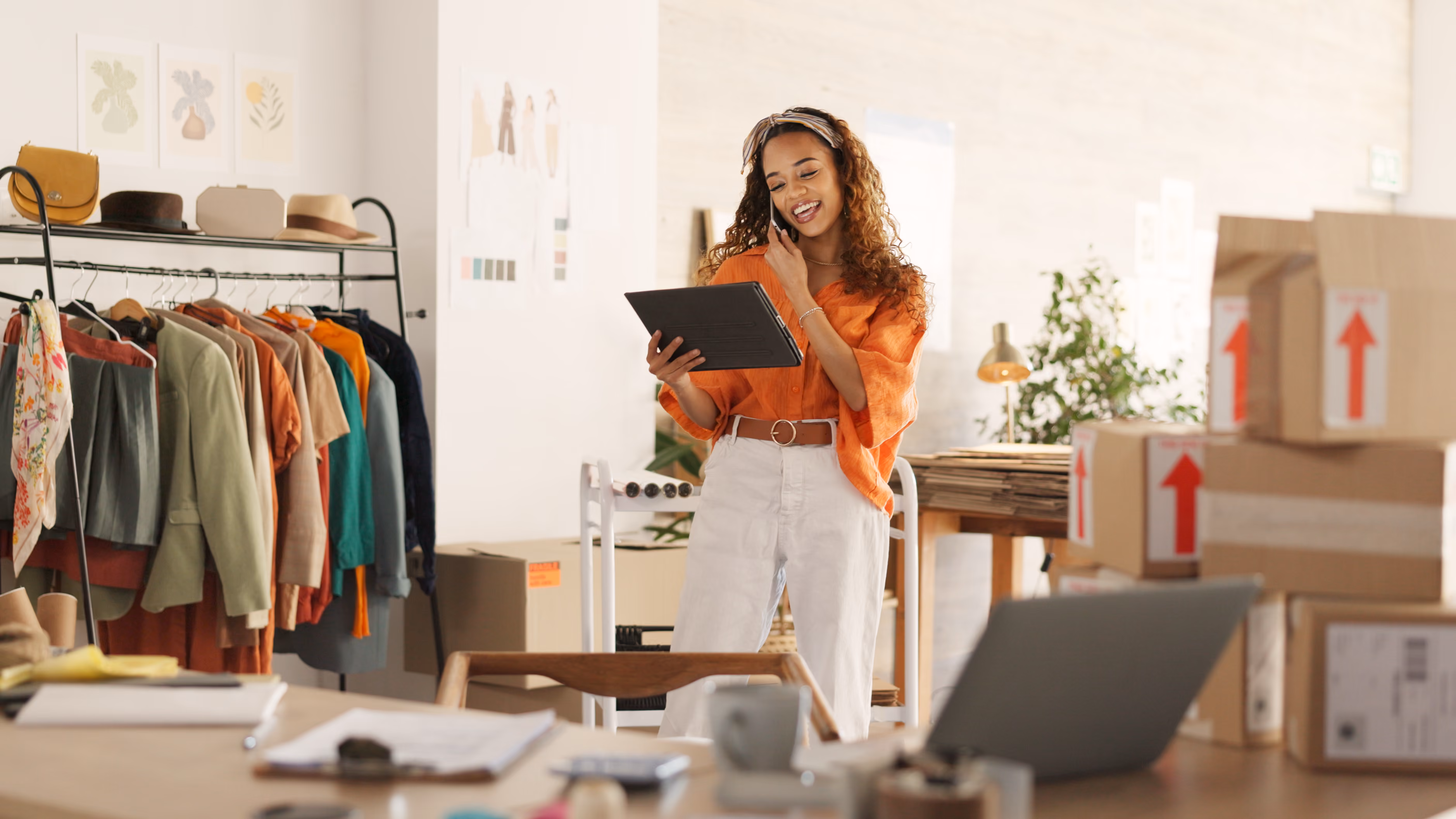 Smiling woman in an orange shirt holding a tablet and talking on a phone in a workspace with clothing and packed boxes.