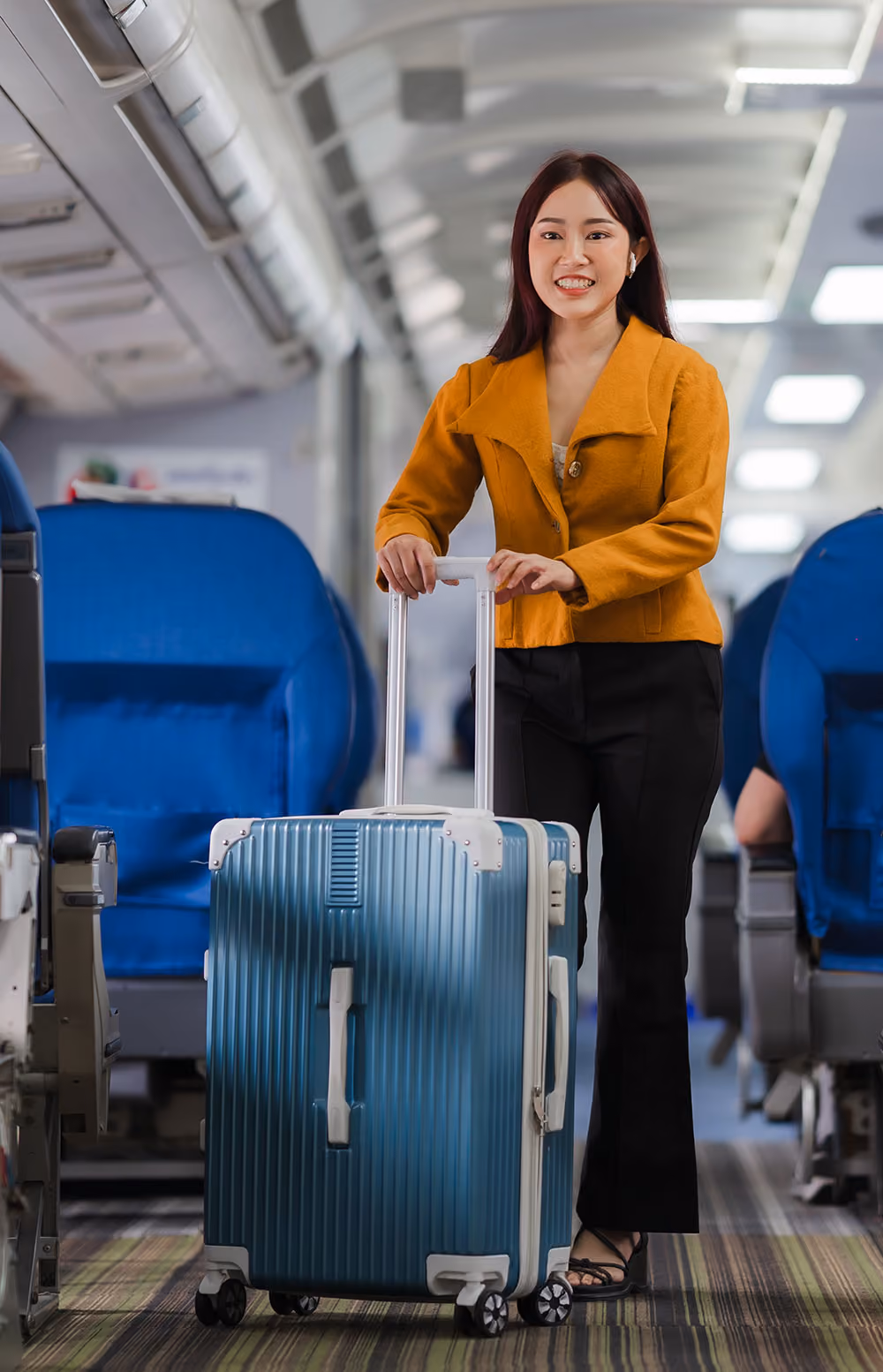 Smiling woman standing inside an airplane aisle with a blue rolling suitcase.