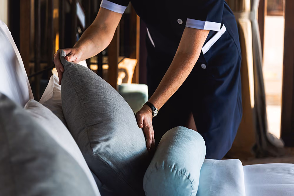 Person in navy uniform adjusting a grey pillow on a white couch in a sunlit room.