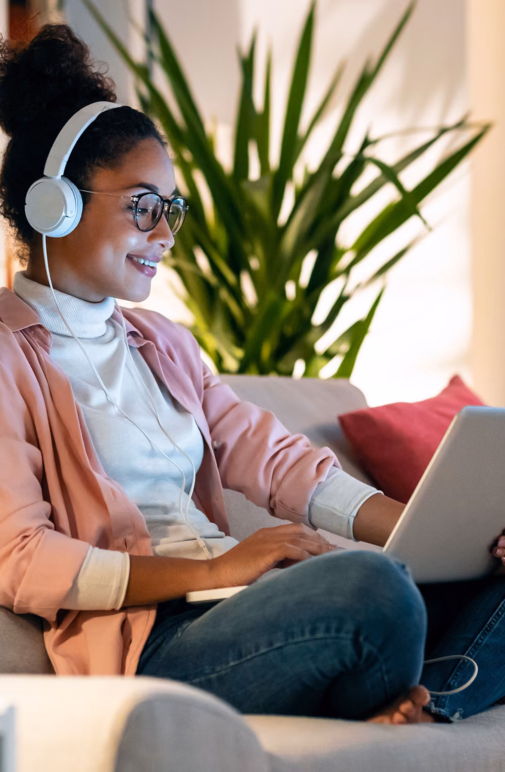 Young woman wearing glasses and headphones sitting on a couch using a tablet, with plants and a red pillow in the background.