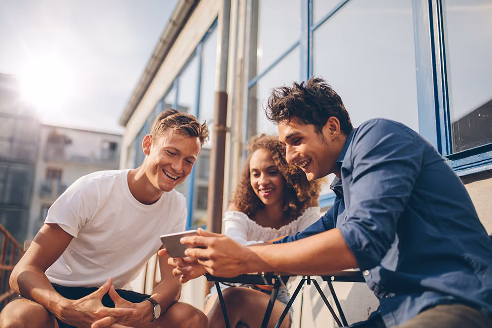 Three young adults sitting outside, smiling and looking at a smartphone together.