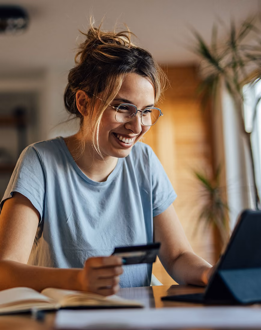 Young woman wearing glasses and a light blue shirt smiling while holding a credit card and using a tablet at a desk.