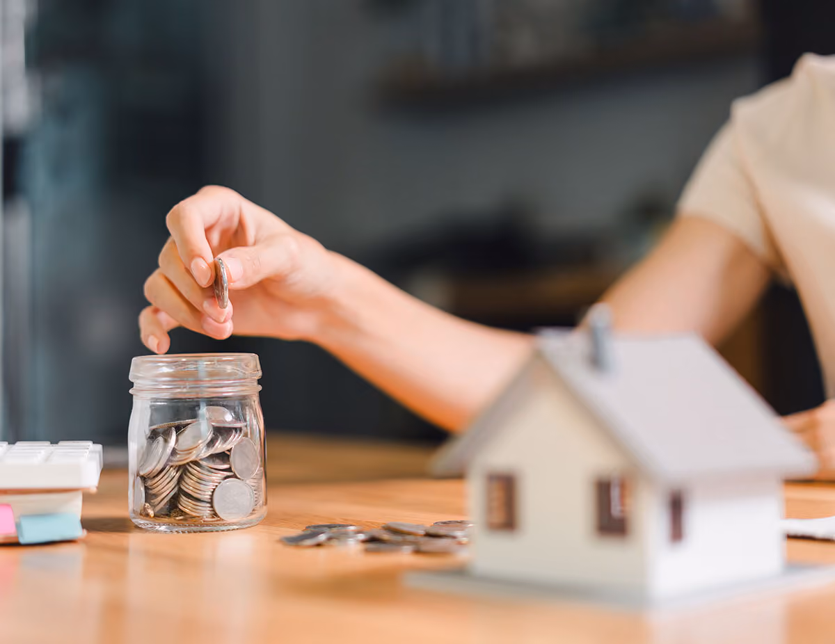 Person placing a coin into a jar filled with coins next to a small model house on a wooden table.