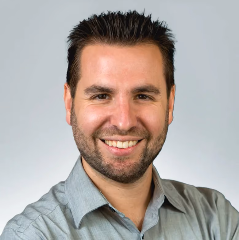 Smiling man with short dark hair and beard wearing a light gray button-up shirt against a plain light background.